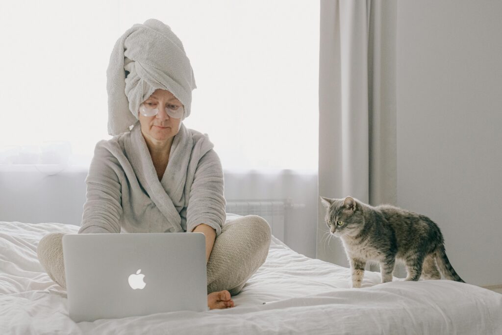 pexels-photo-3846135-3846135 A woman in a robe uses a laptop while a cat sits beside her in a cozy bedroom setting.